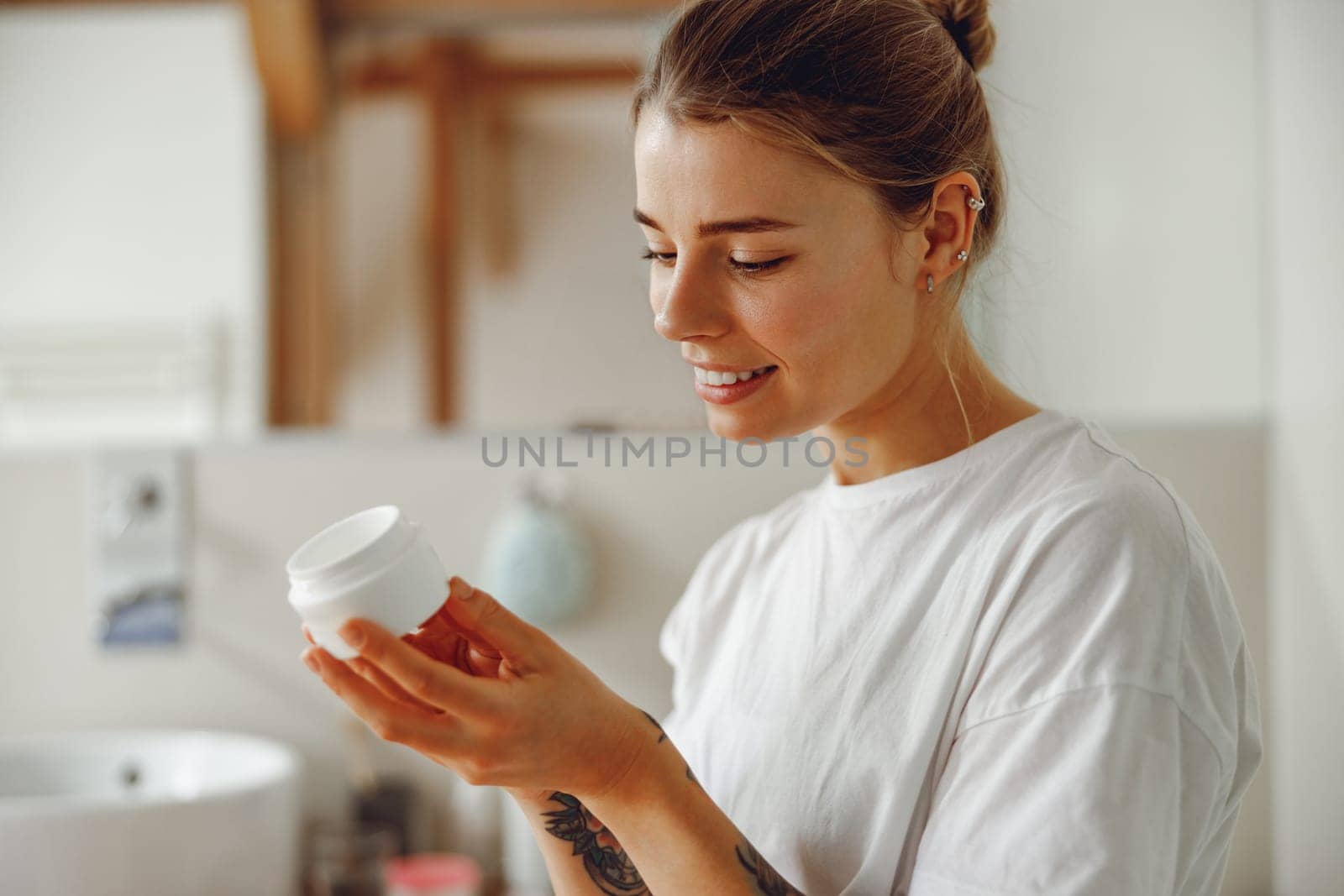 Smiling woman looking on jar with skincare cream sitting in bathroom. Home beauty routine by Yaroslav_astakhov