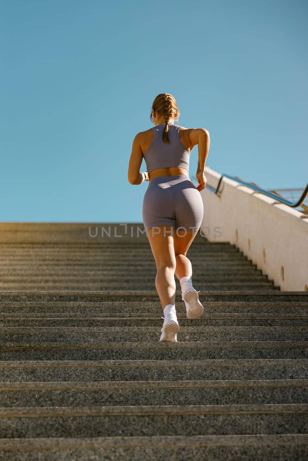 Back view of active female athlete in sportswear running on steps outdoors on a sunny day by Yaroslav_astakhov