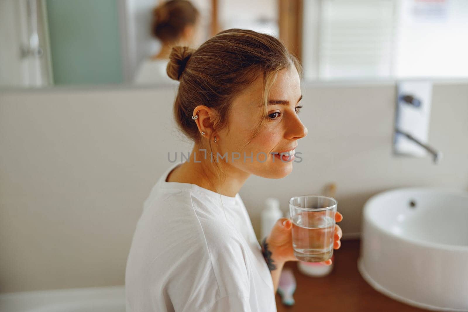 Smiling young woman drinking glass of water in the bathroom. Morning beauty routine