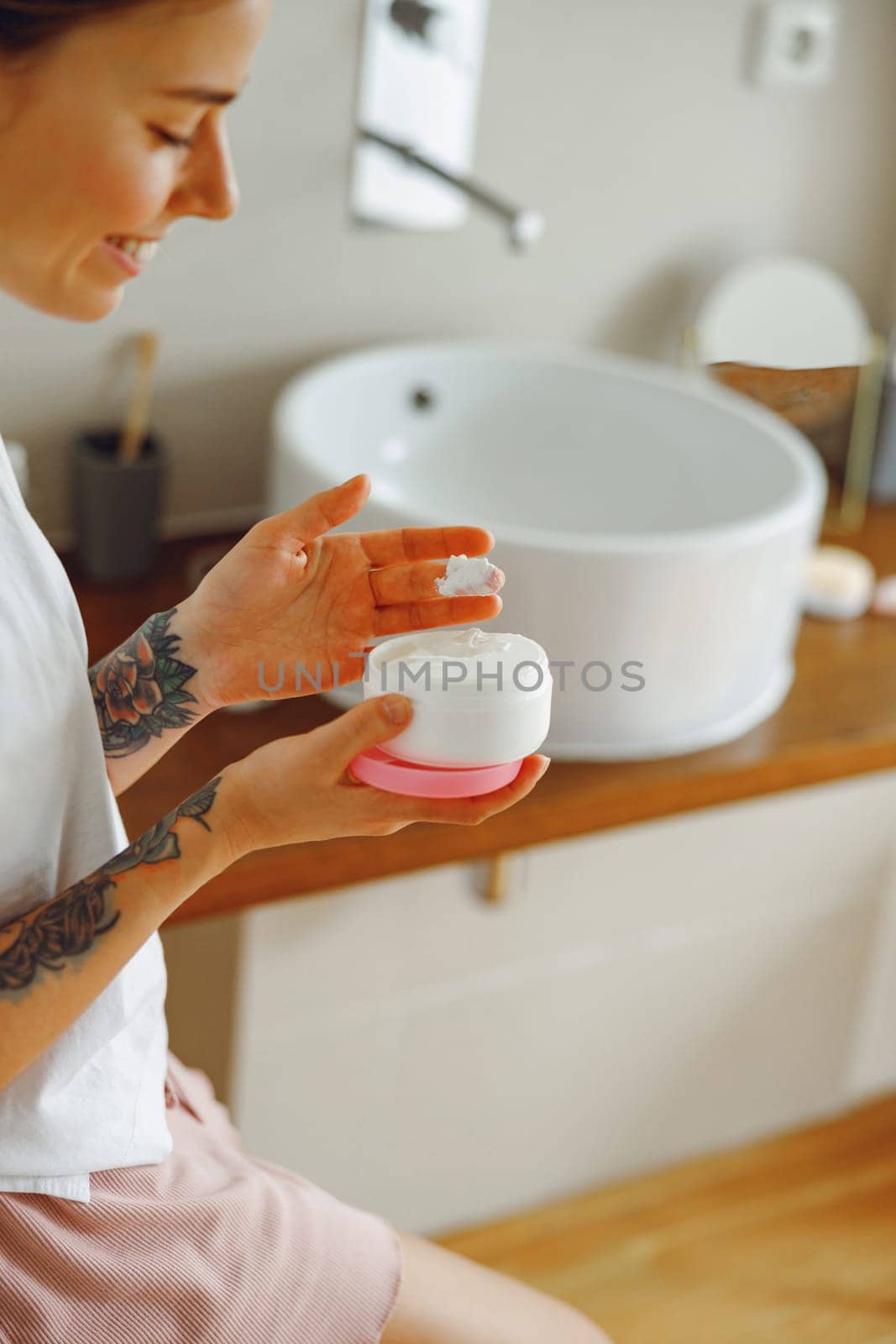Smiling young woman use jar with face cream while standing in bathroom. Home beauty routine by Yaroslav_astakhov