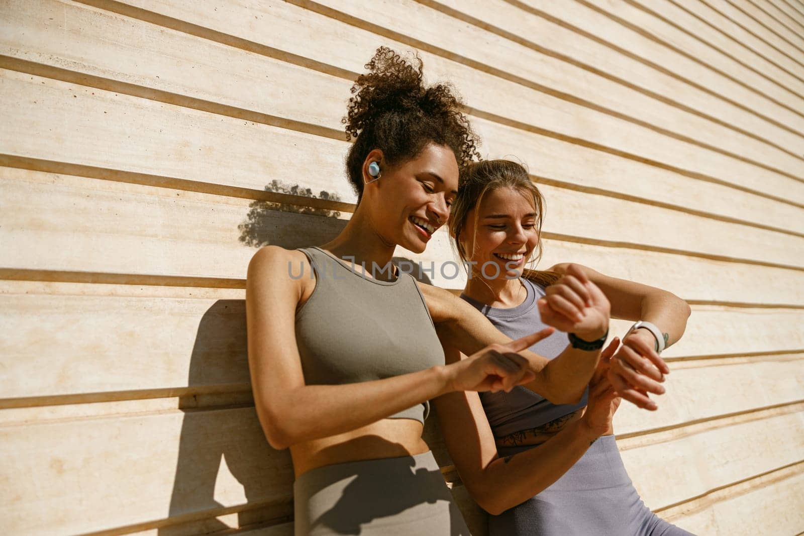 Smiling woman with female friend looking on smartwatch before exercising standing near wall outdoors by Yaroslav_astakhov