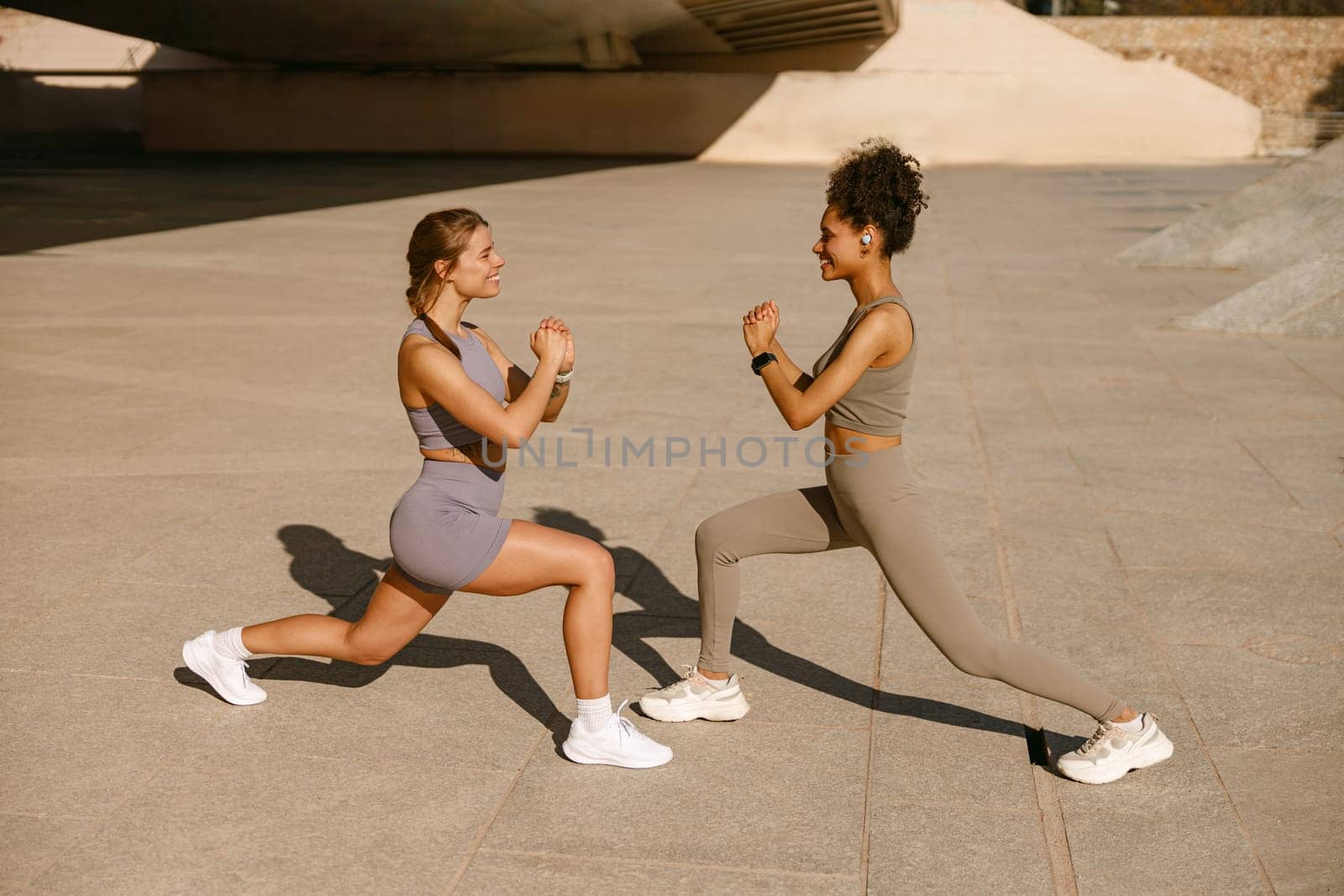 Young sportswomen in tracksuits are stretches legs outside on modern building background