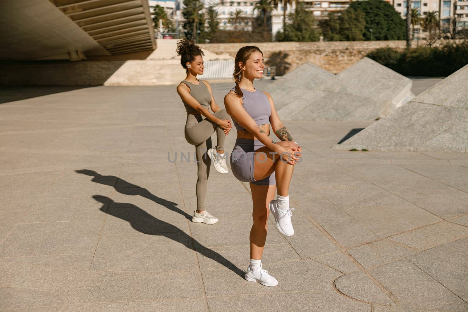 Two young sporty women in tracksuits are stretches legs outside on modern building background by Yaroslav_astakhov