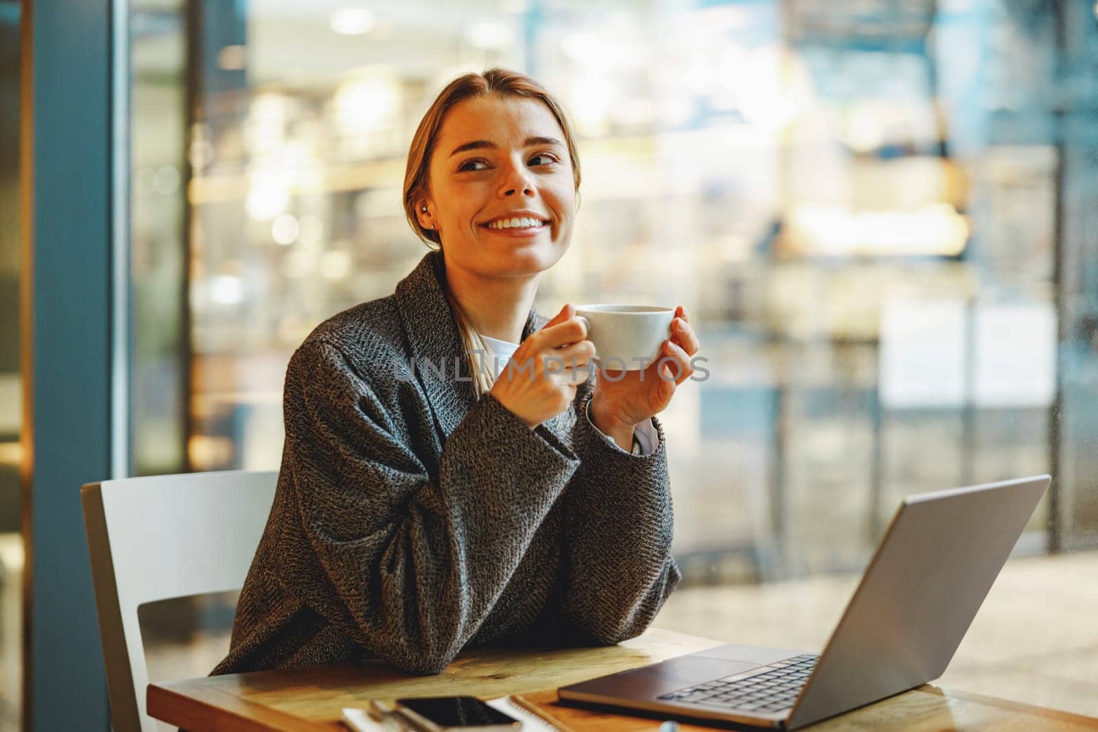 Stylish female freelancer is drinking coffee in cafe while working on laptop near window by Yaroslav_astakhov