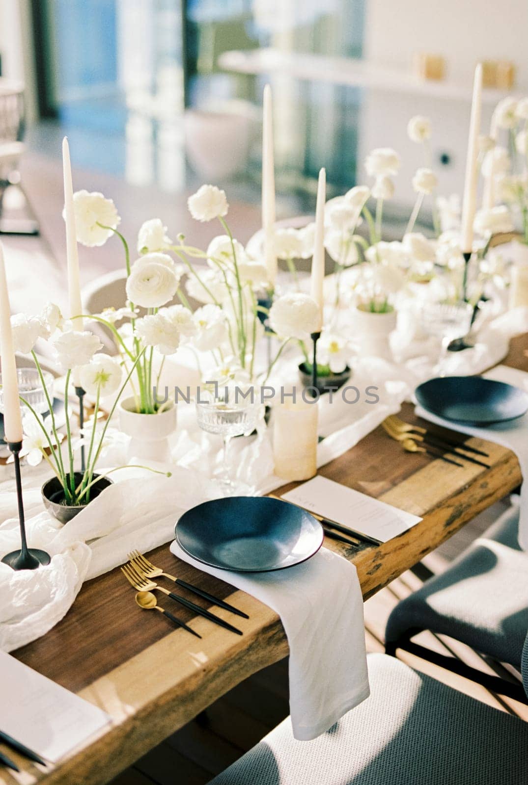 Black plates stand on a wooden table near white bouquets of flowers and candles on a narrow tablecloth by Nadtochiy