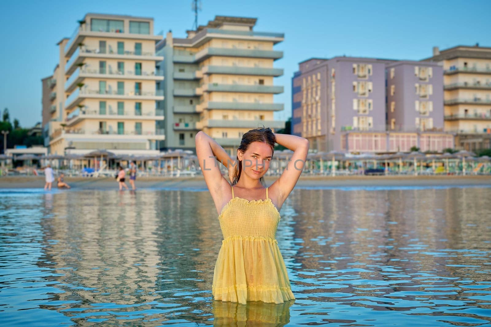 With her hands raised, a young woman early in the morning, against the backdrop of city buildings, goes into the water on the city beach, watching the sunrise