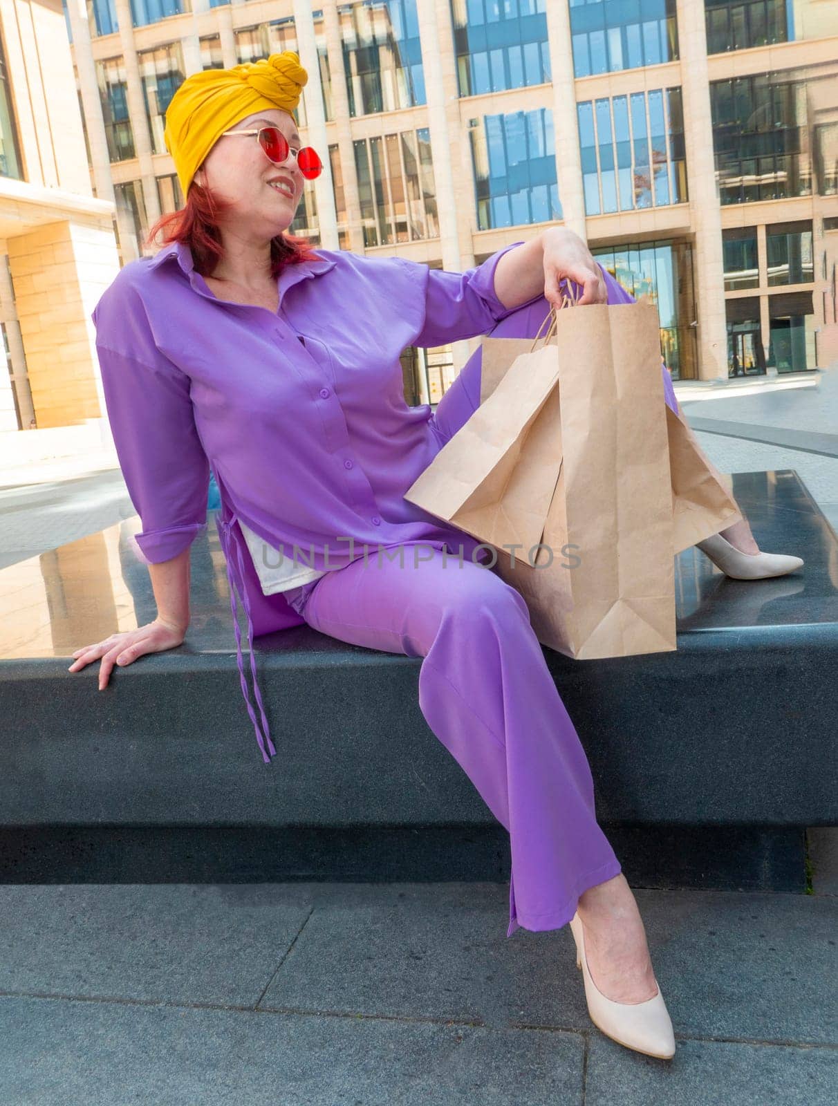 woman with craft shopping bags in the city. The concept of fashionable Consumerism and ecology. An adult Asian woman in a purple suit with red and yellow accents enjoys shopping . a sunny day.