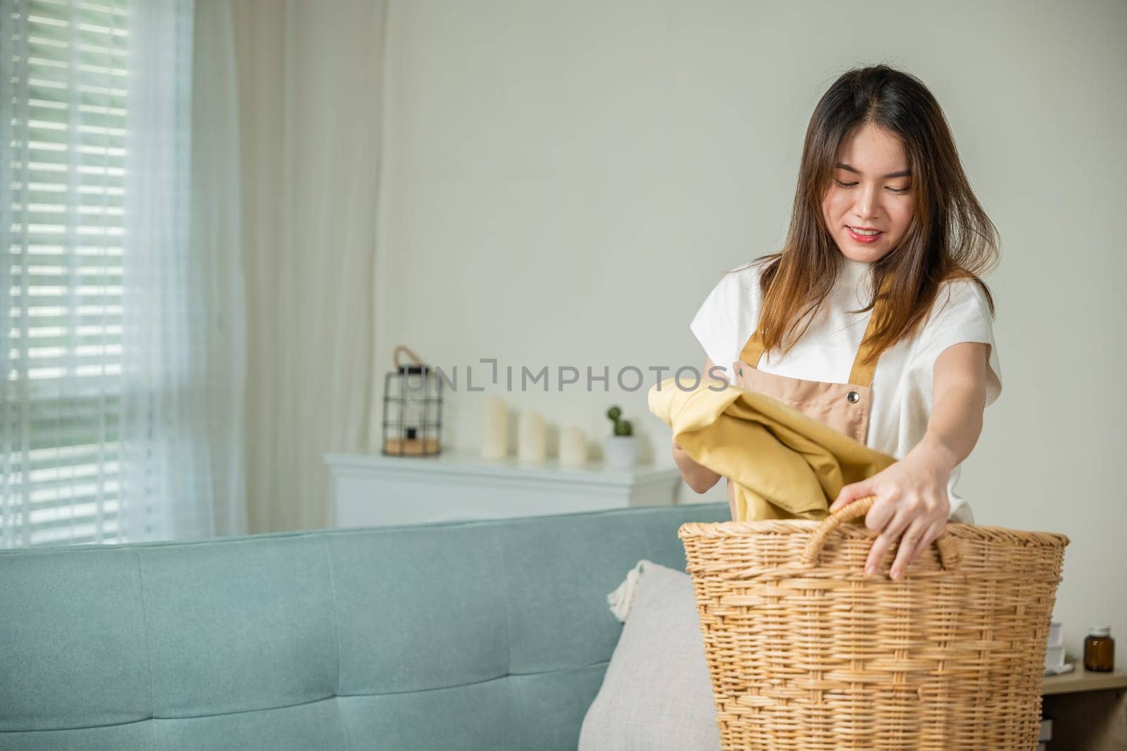 Beautiful housekeeper doing housework holds wooden basket of clean messy dirty clothes to washing at home, Asian housewife woman holding basket with heap of different clothes on sofa in living room