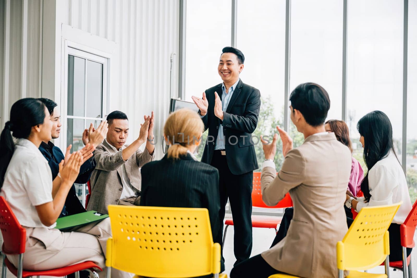 Asian man presents his work in meeting room receiving praise and compliments from colleagues. The audience claps with happiness celebrating his achievement. It signifies teamwork and business success.