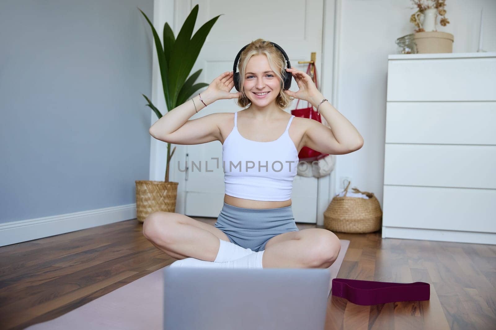 Portrait of cute young fitness woman, connects to online training session via laptop, wearing wireless headphones during workout, sits at home on rubber yoga mat by Benzoix