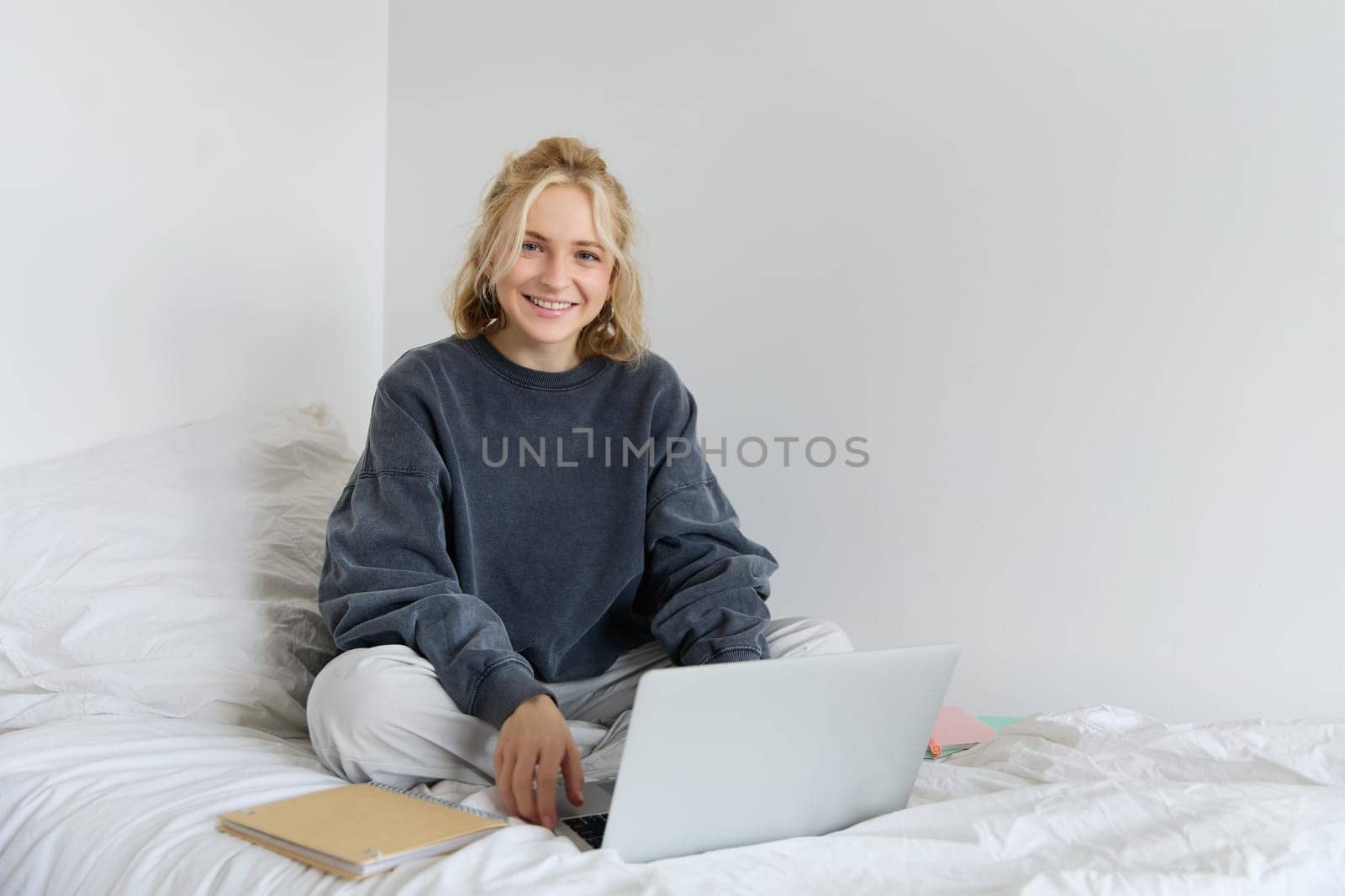 Portrait of young smiling woman studying in her bed, working from home in bedroom, sitting with laptop and notebooks on lotus pose, looking happy and relaxed.