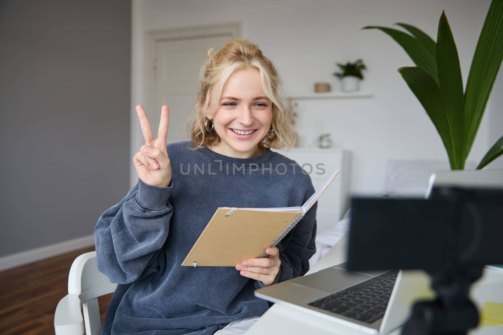 Image of smiling girl records video of herself on digital camera, shows peace sign, sits in front of laptop, holds notebook.