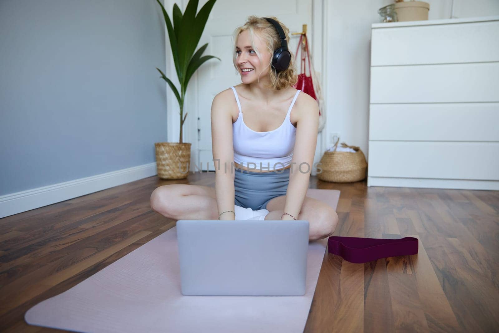 Portrait of smiling sporty woman at home, doing workout on rubber yoga mat, using laptop and wireless headphones by Benzoix
