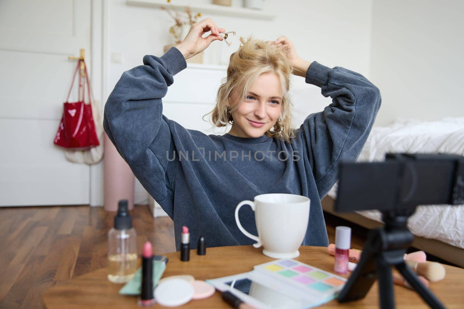 Lifestyle blogger records video of herself while doing hairstyle, showing makeup lifehacks for social media followers, vlogging, using camera for her blog, sitting in a room, drinking tea.