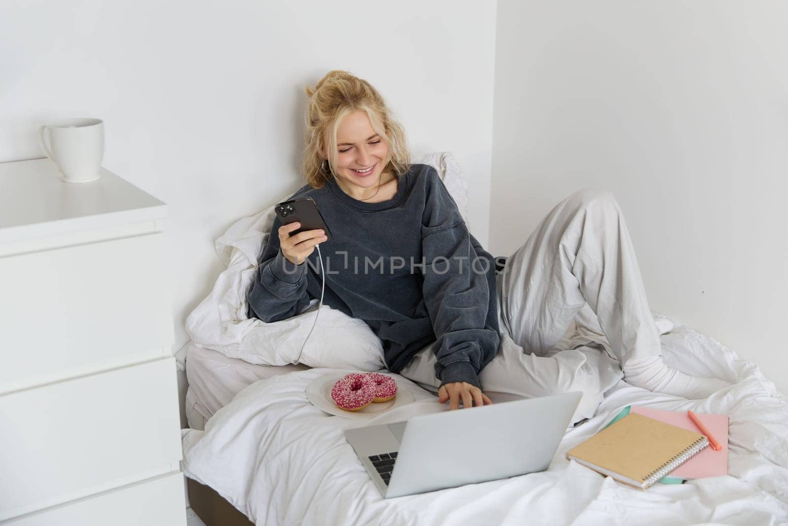 Portrait of smiling candid woman, lying in bed with doughnut, using smartphone and laptop, resting at home in bedroom, watching tv show or chatting online.