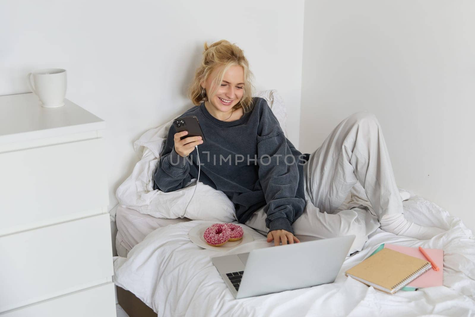 Lifestyle and people concept. Young beautiful woman, staying at home, lying in bed with laptop and smartphone, eating doughnut, enjoying free time, spending weekend at home, watching movie online.