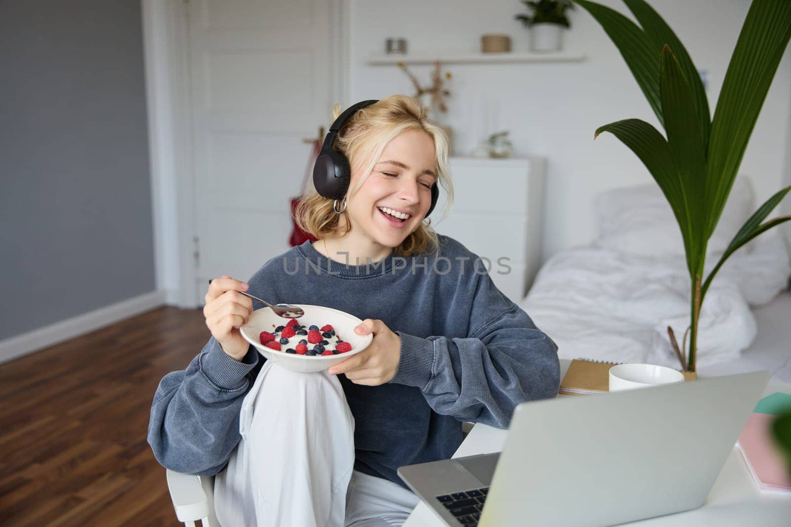 Portrait of smiling young woman, watching tv show in headphones, eating breakfast and looking at laptop screen.