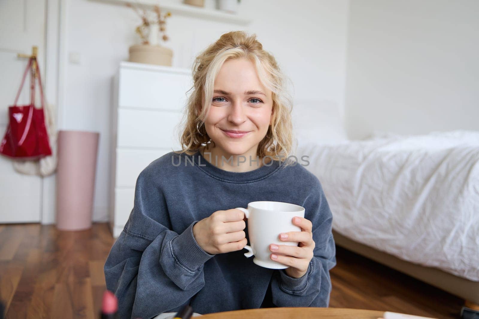 Image of young teenage girl sitting in her bedroom on floor, drinking cup of tea and enjoying day at home, smiling and looking at camera.
