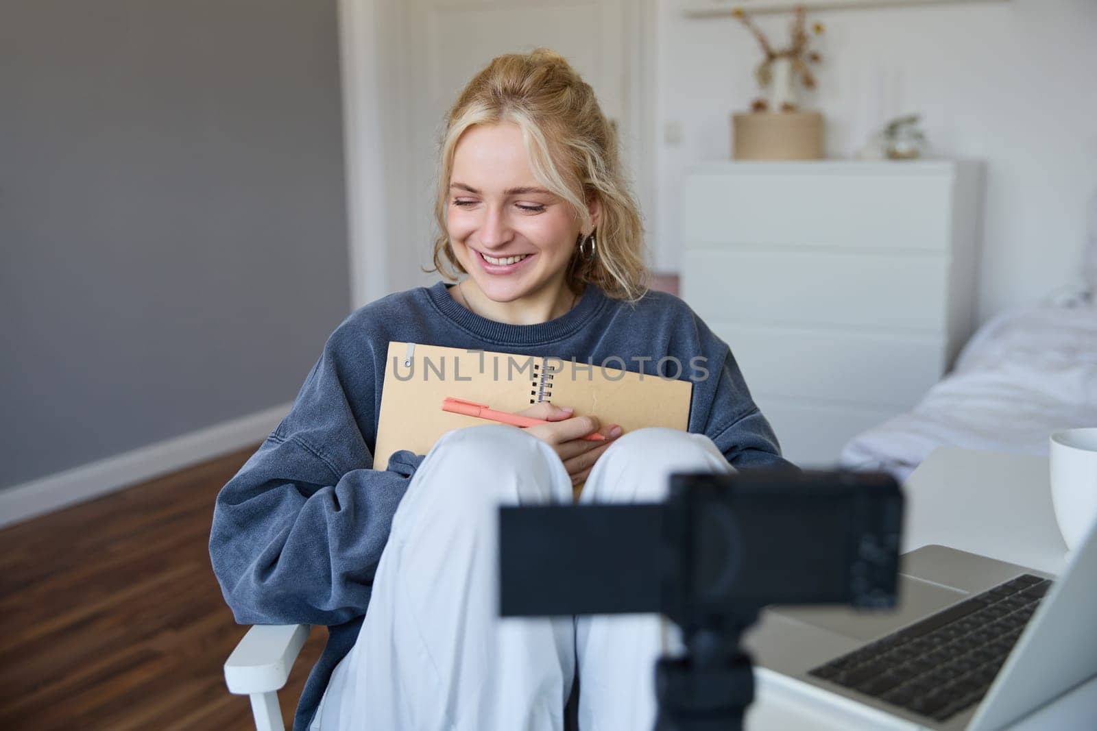 Portrait of smiling cute woman, lifestyle blogger, sits in her room with daily journal or planner, records video on digital camera, creates content for social media about daily routine.