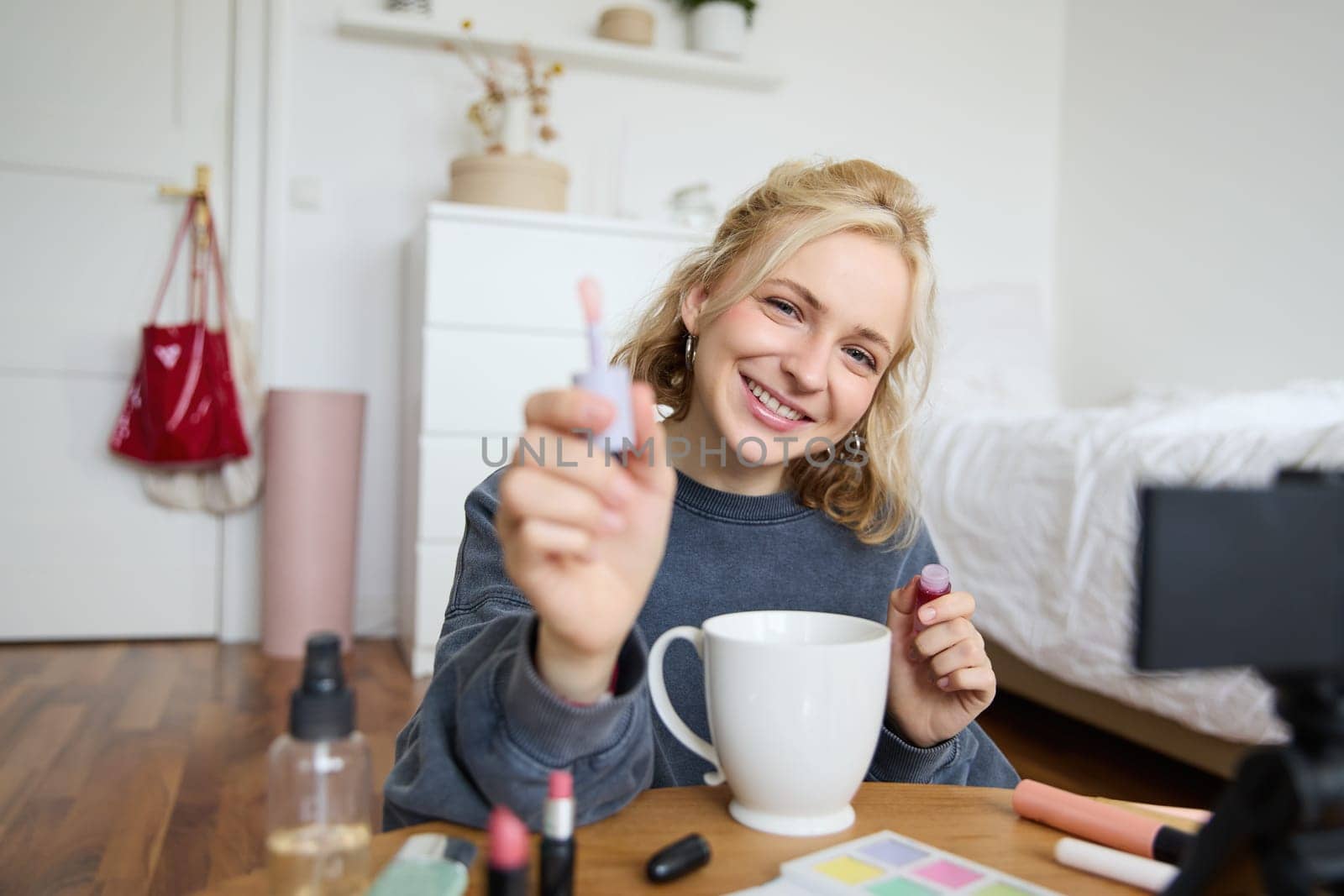 Portrait of smiling woman shows lip gloss, advertising makeup on her social media account, recording video on digital camera, content maker working on new project in her room.