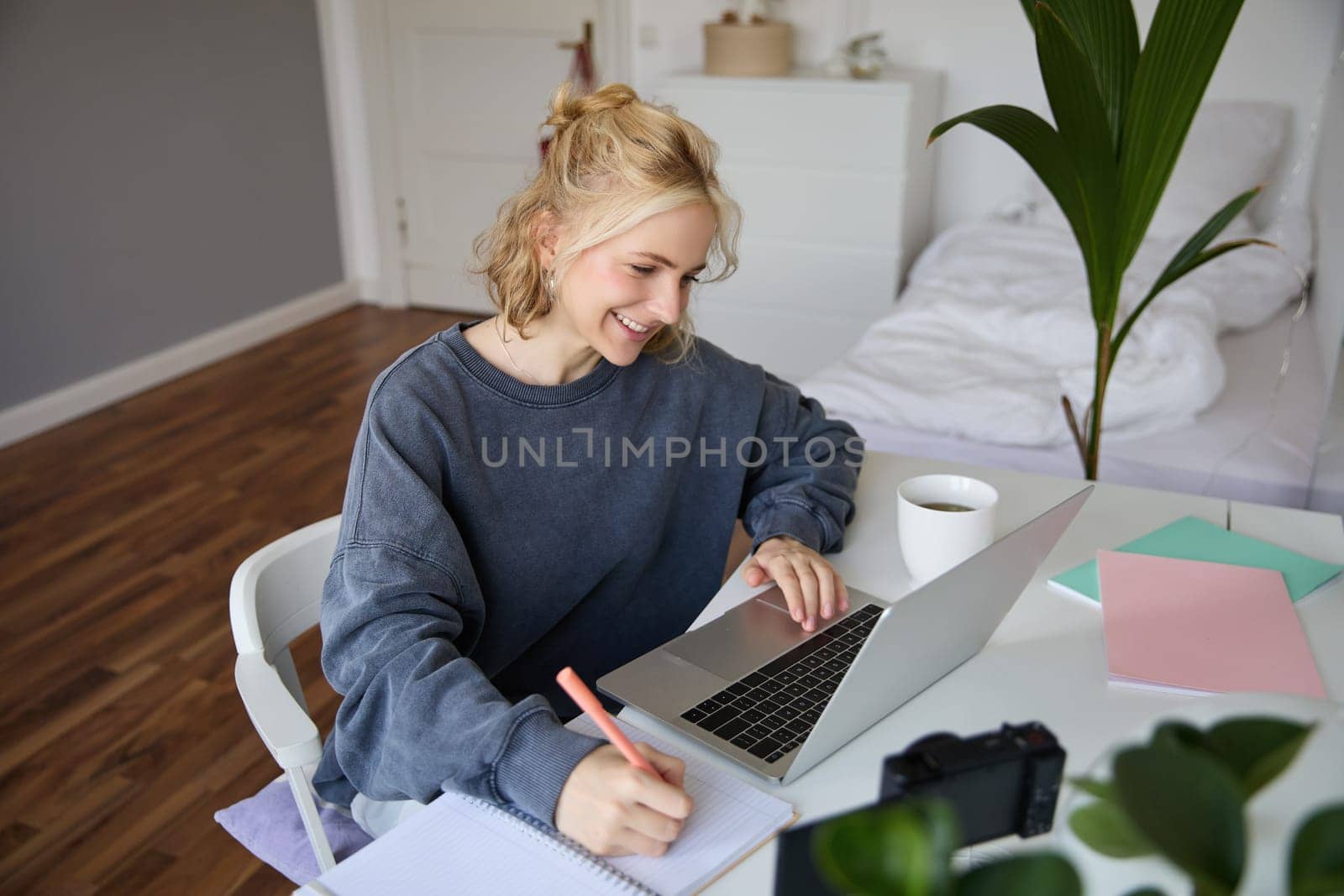 Portrait of smiling young woman, female student, doing distance learning course, using laptop, studying at home, writing down, making notes.