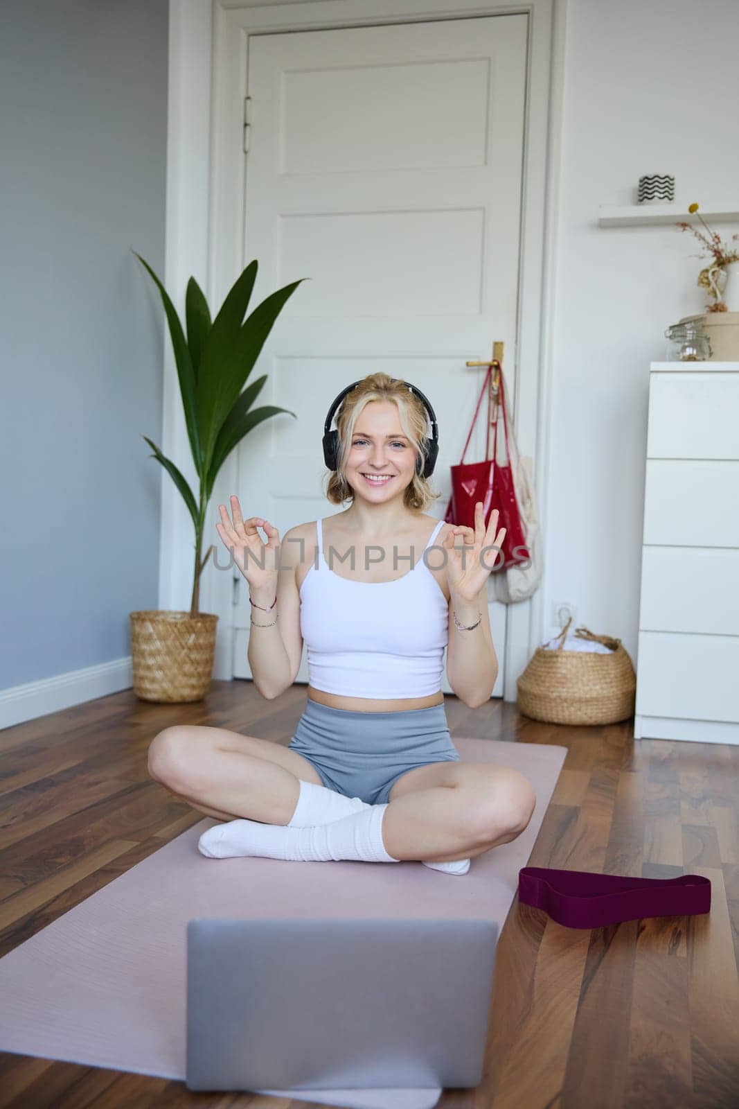 Vertical shot of smiling woman in wireless headphones, fitness instructor connects to online workout training session, shows exercises using laptop camera, sits on rubber yoga mat at home by Benzoix