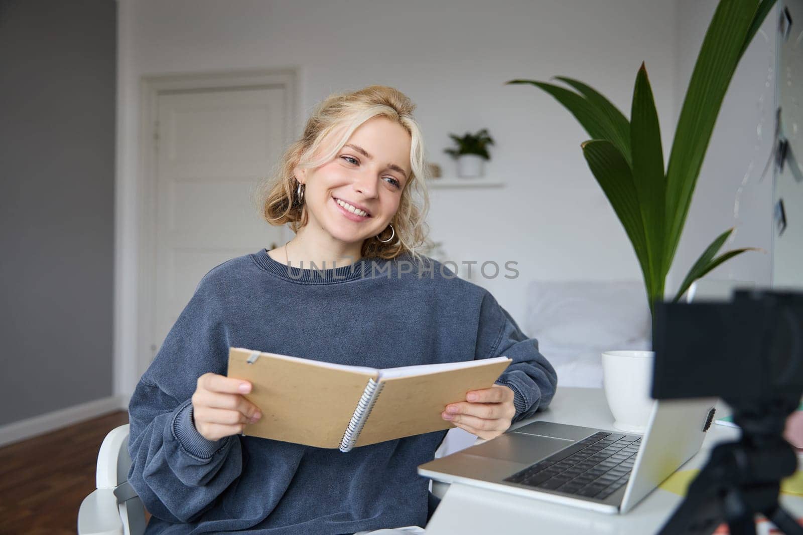 Portrait of smiling blond woman, sitting in bedroom, using laptop and digital camera, recording video for lifestyle blog, reading, using her notebook.