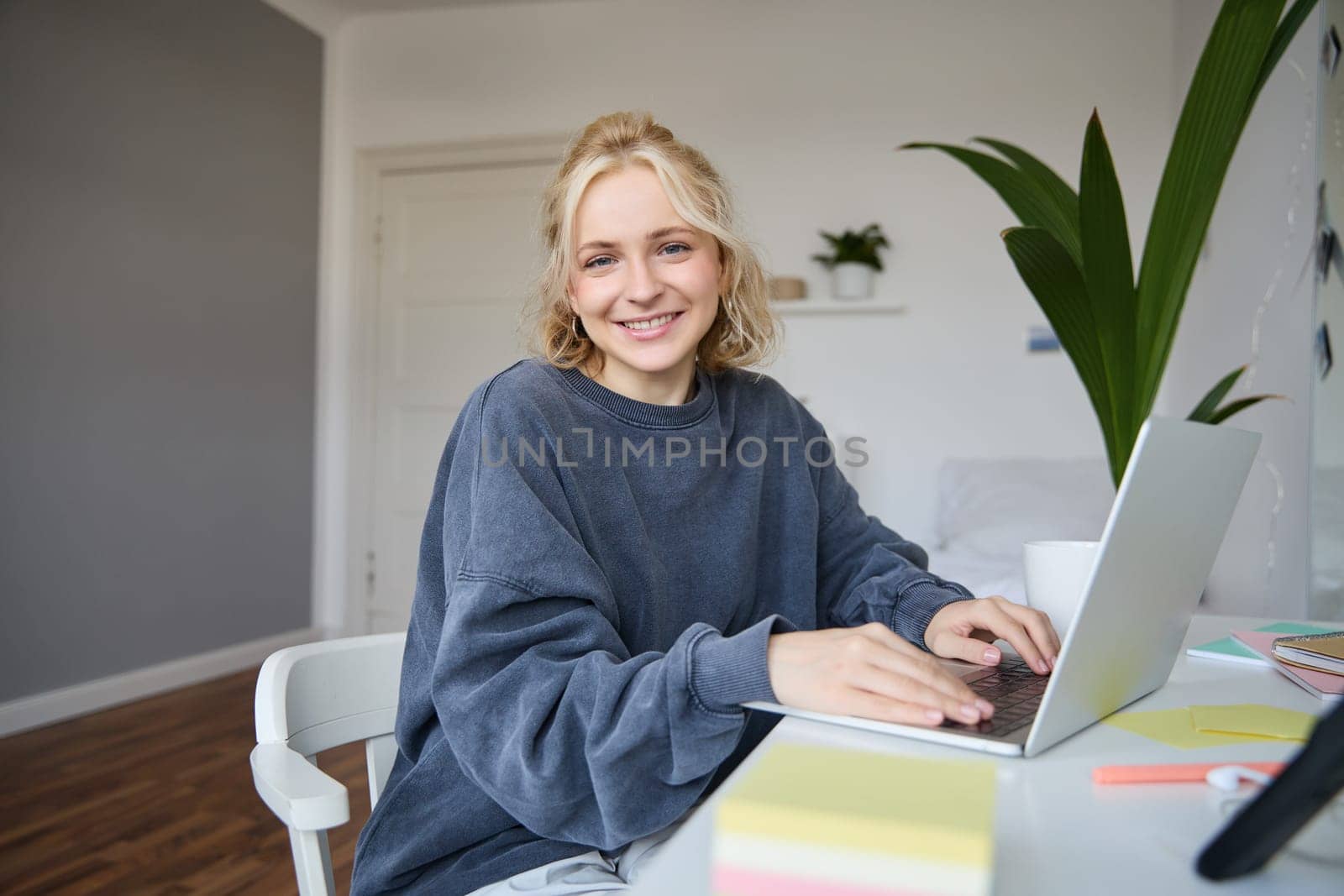Portrait of smiling young woman, college student sits in her room, does homework, studies remotely from home, uses laptop for freelance work.