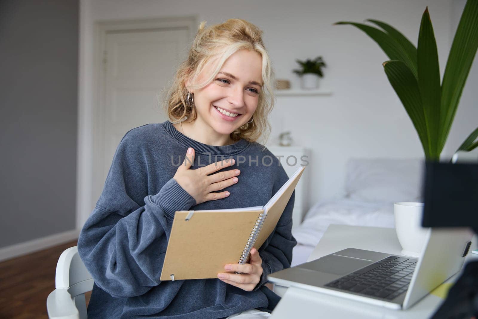 Portrait of young smiling blond woman, working from home, online chatting, using digital video camera, recording vlog, holding notebook, reading notes, explaining something.