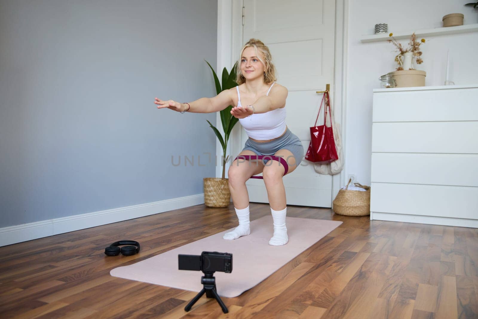 Portrait of woman, fitness instructor at home, recording video about workout, showing how to do leg exercises, squats with elastic resistance band, working out indoors on yoga mat.