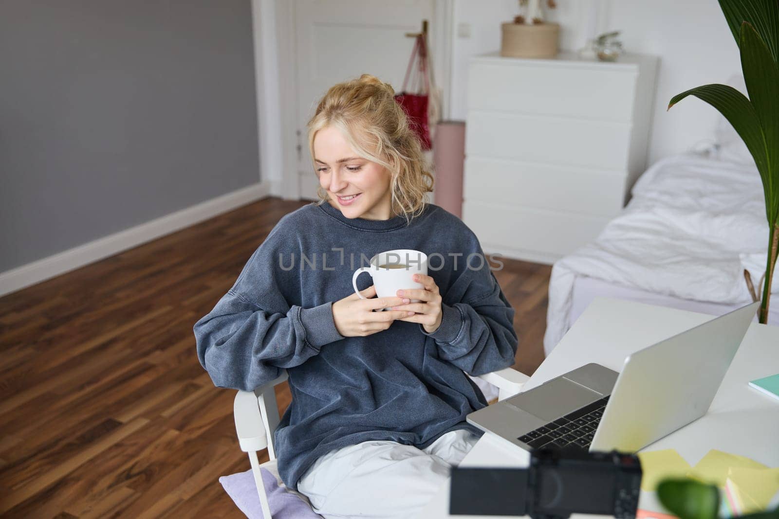 Portrait of young smiling woman, vlogger recording lifestyle video in her room, sits in front of laptop and digital camera, drinks tea, creates content for social media account.