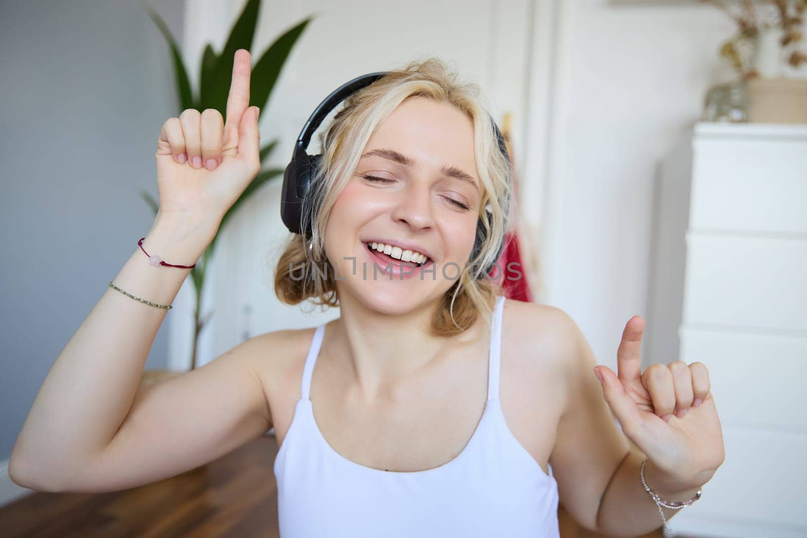 Portrait of happy and carefree blond woman, listens to music in wireless headphones, dancing and singing along favourite song, spending time alone at home by Benzoix