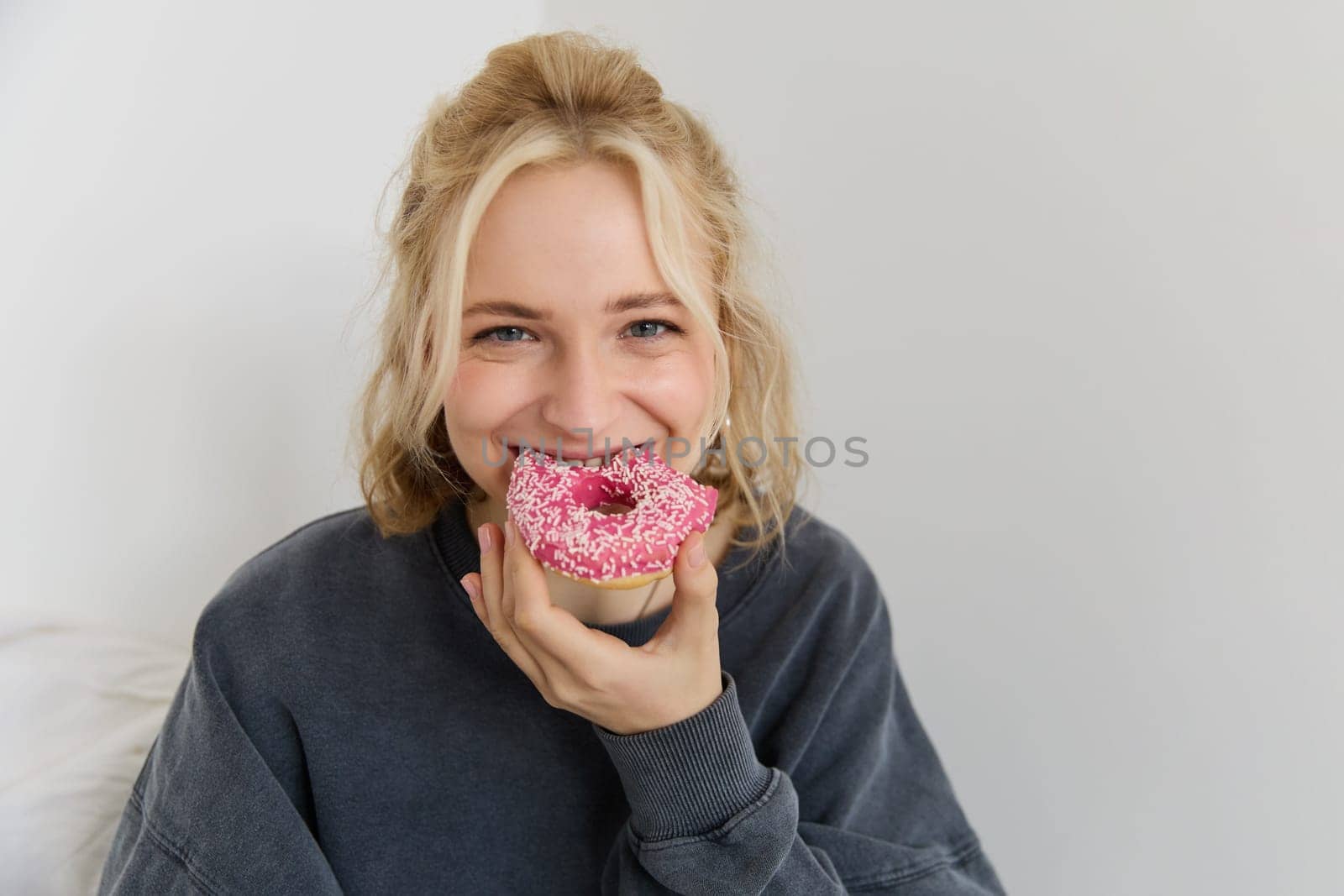 Portrait of cute blond girl holding pink doughnut with sprinkles on top, showing her favourite food.