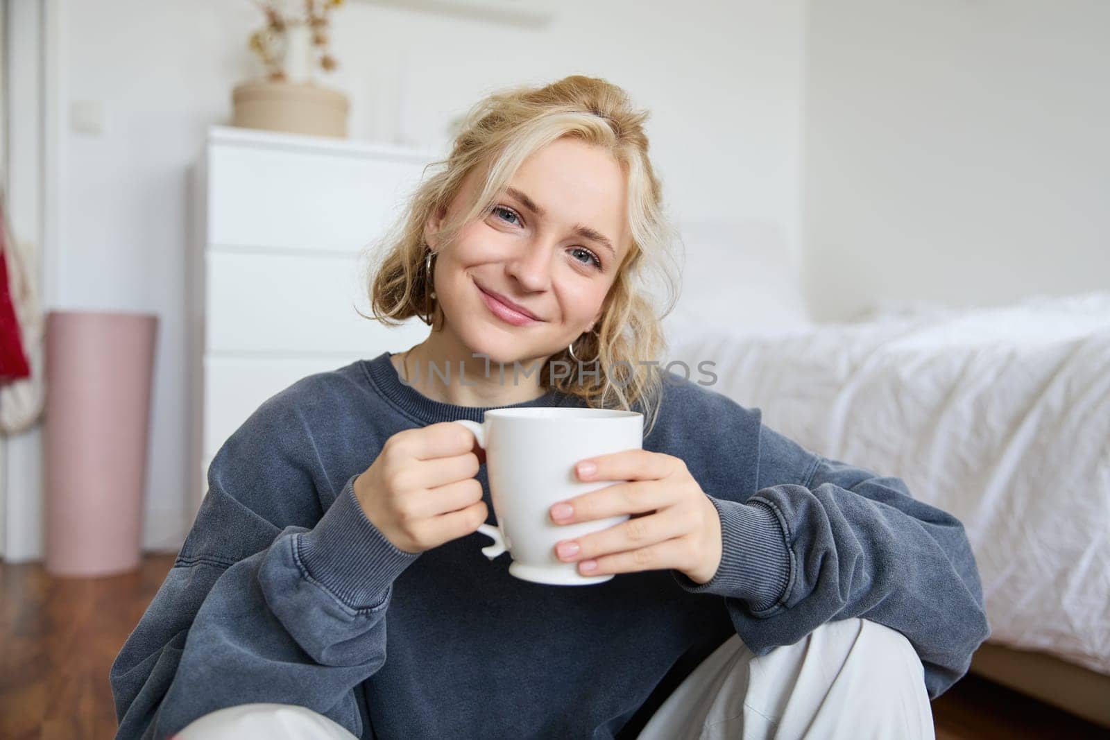 Image of young teenage girl sitting in her bedroom on floor, drinking cup of tea and enjoying day at home, smiling and looking at camera.