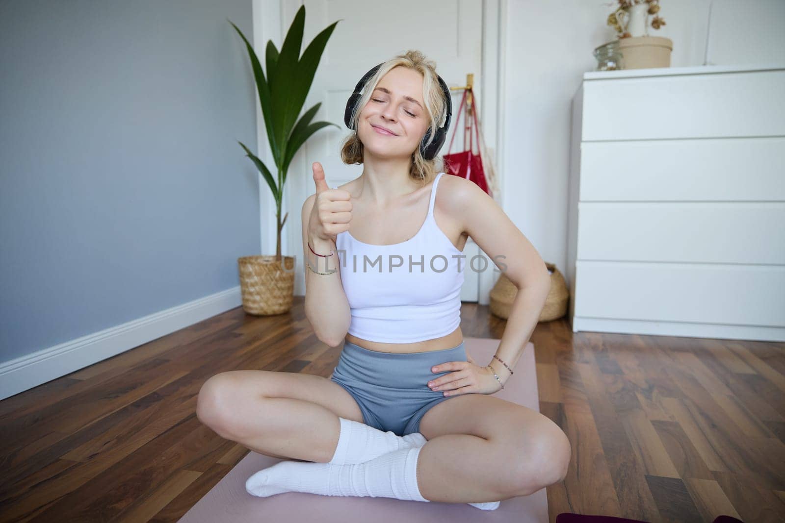 Portrait of happy, fit and healthy young woman sitting on yoga mat, wearing wireless headphones, showing thumbs up with satisfied face, pleased with workout training at home.
