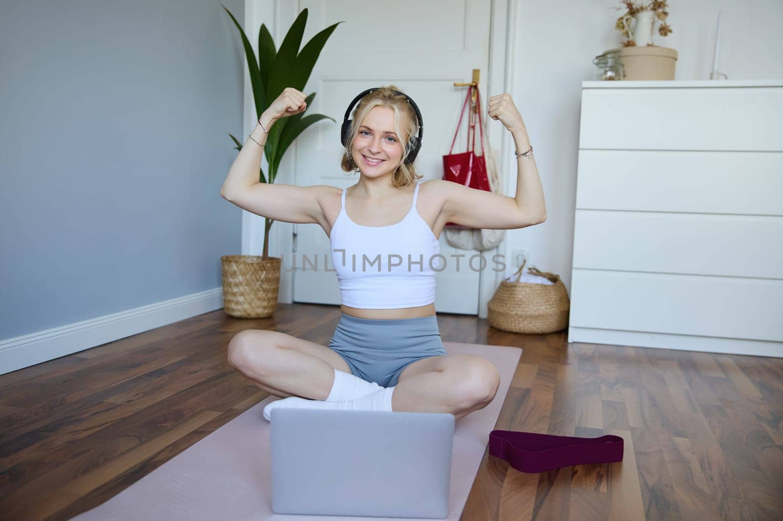 Portrait of young happy woman, fitness blogger in wireless headphones, working out at home, sits on rubber mat with laptop and wireless headphones, shows her muscles.