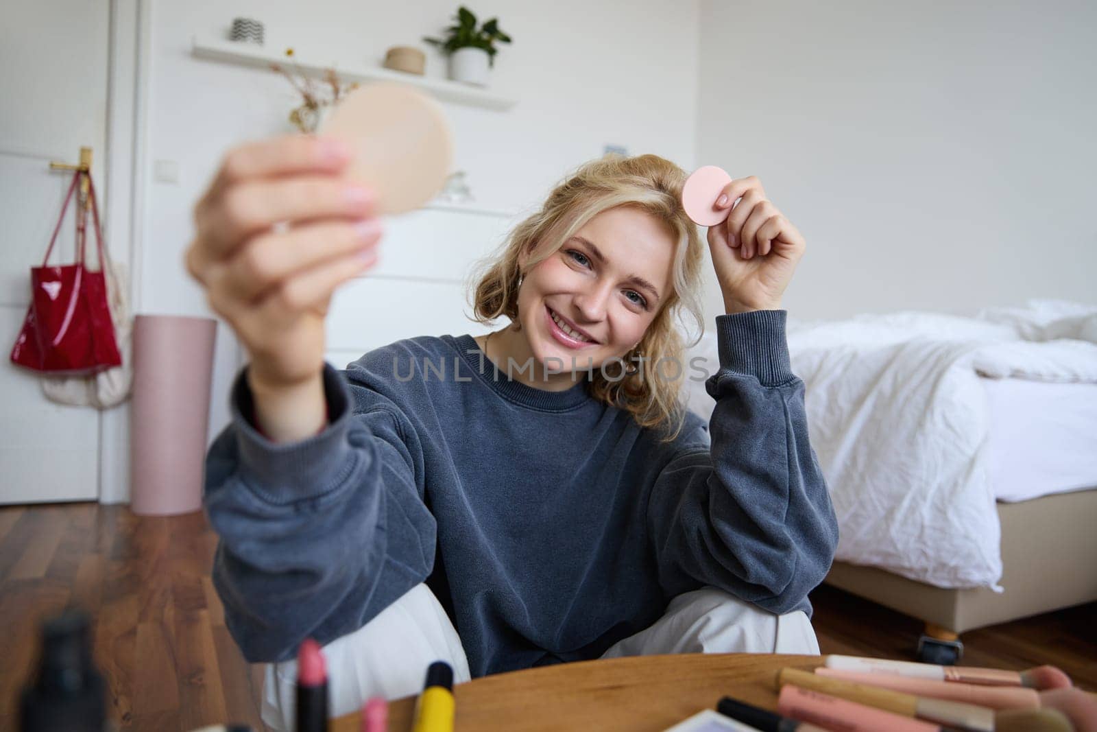 Portrait of young woman, content creator, showing beauty makeup products at camera, sitting on floor in bedroom and smiling.