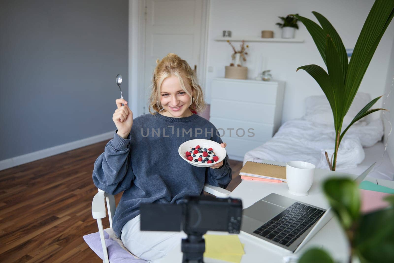 Portrait of cute young girl vlogger, showing her breakfast on camera, recording vlog in her bedroom, holding dessert, eating.