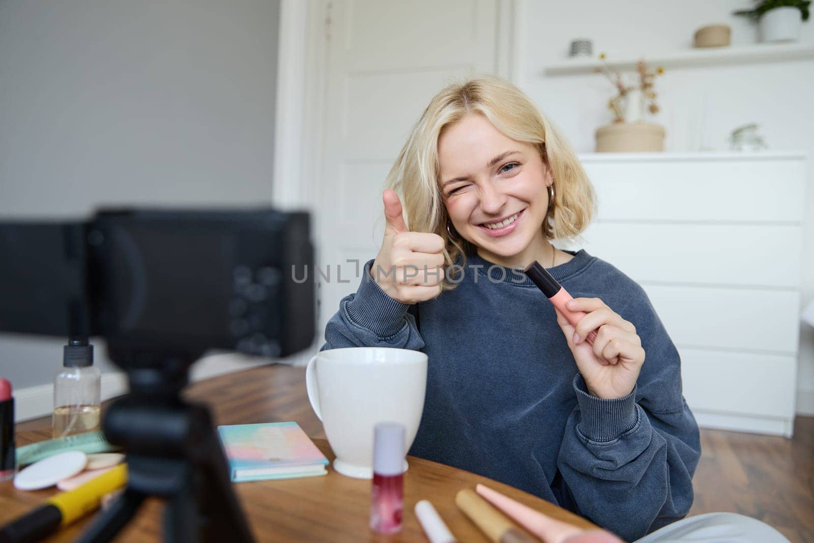 Portrait of smiling, beautiful young woman, lifestyle blogger recording a video in her room, sitting in front of camera on stabiliser, showing thumbs up and wink face, recommending mascara.