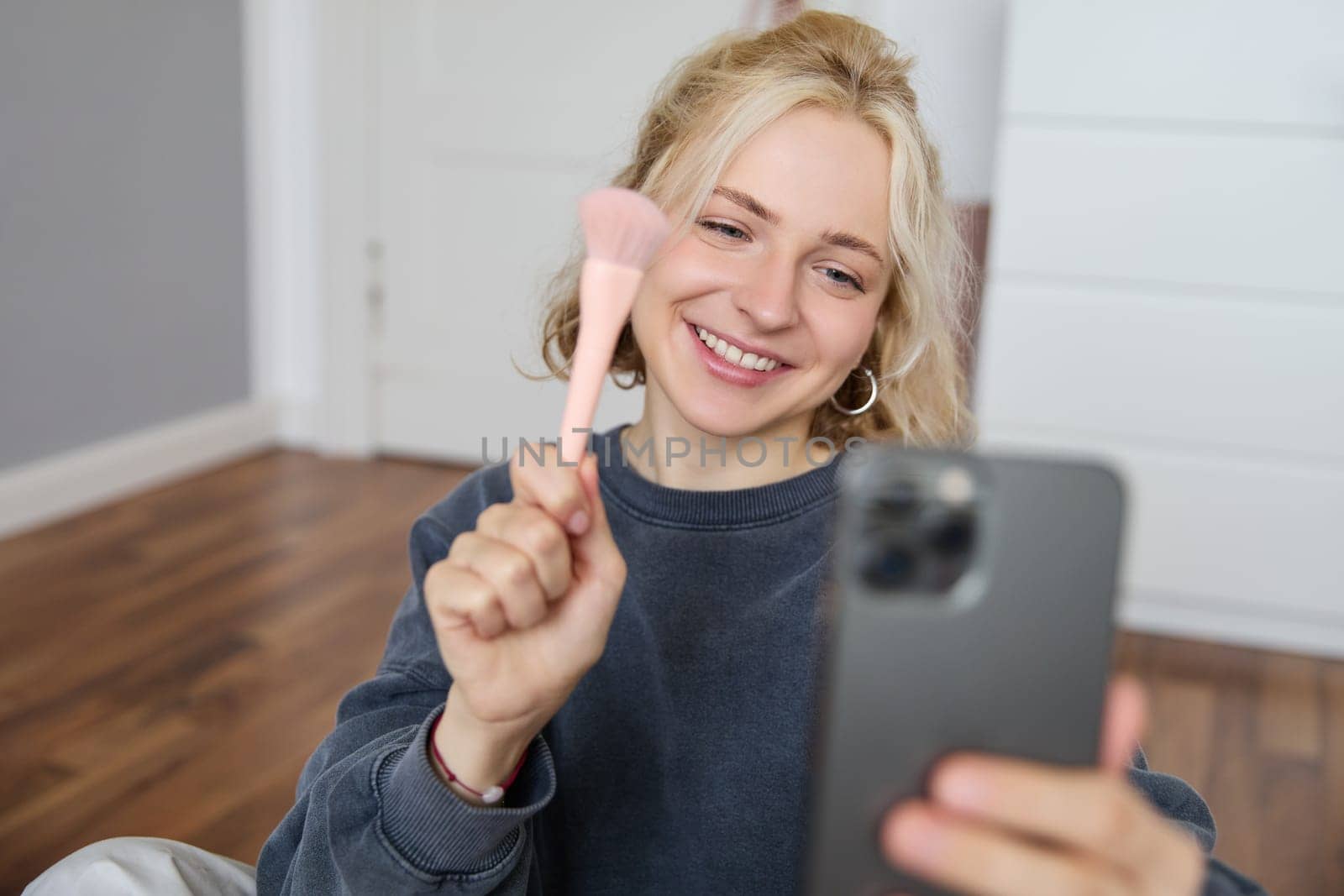 Portrait of young woman, social media influencer, taking selfies in her room, sitting on floor, holding smartphone and posing for a photo.