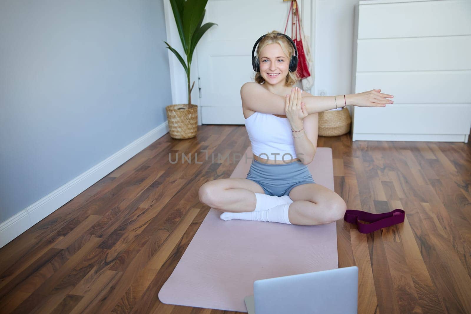 Wellbeing and fitness concept. Woman in headphones, sits on yoga mat, watches workout video on laptop, stretches arms, does exercises.