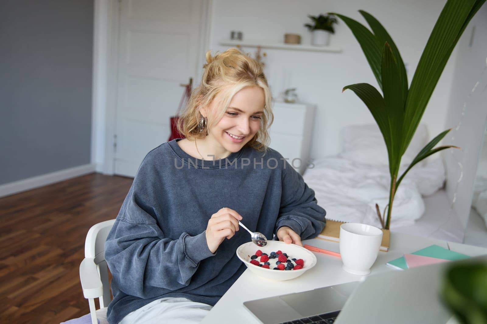 Portrait of smiling blond young woman, eating in front of laptop, watching videos online while having breakfast, enjoying dessert, sitting in bedroom.