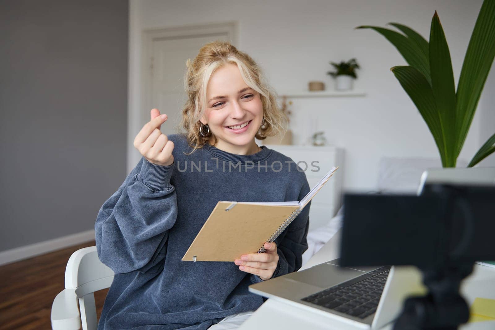 Cute young woman in her room, holds notebook, sits with laptop, looks at digital camera, records vlog or online tutorial.