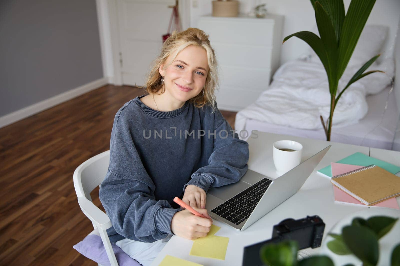 Portrait of smiling young woman, female student, doing distance learning course, using laptop, studying at home, writing down, making notes.