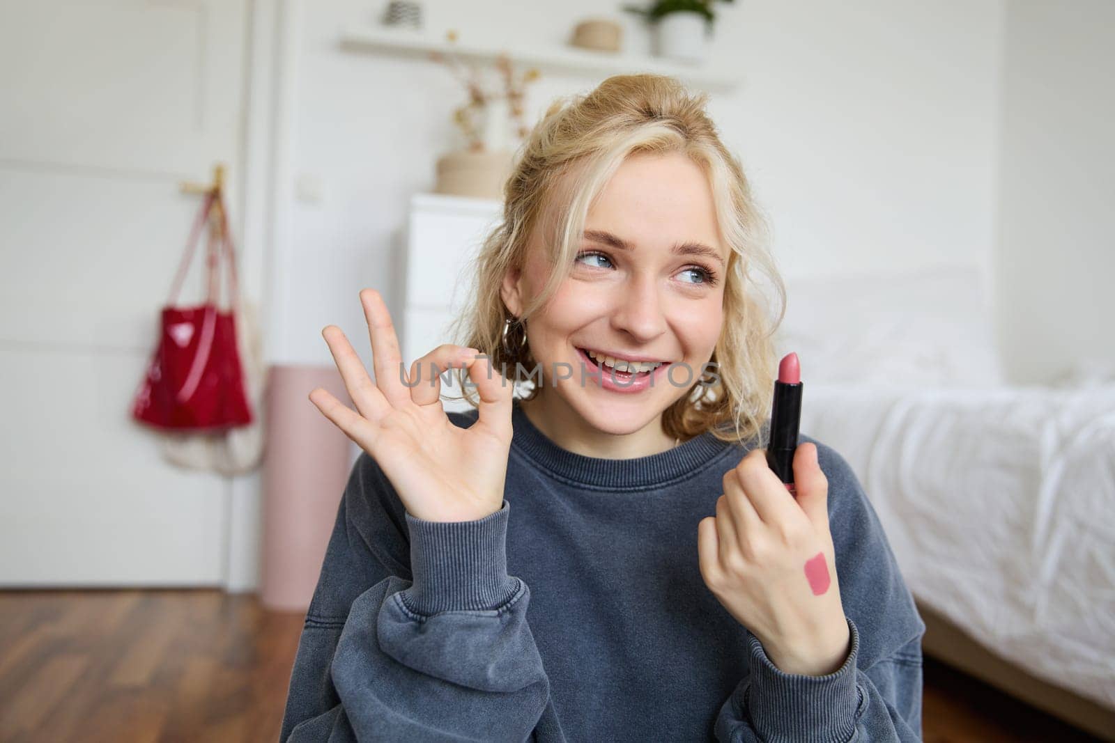 Young female blogger, content creator showing lipstick and okay hand sign, recommending beauty product for her audience on social media, recording vlog in room.