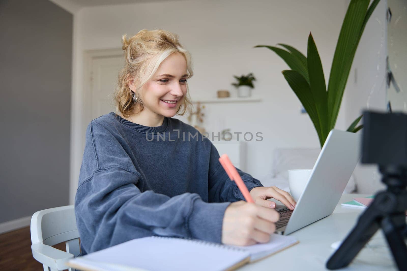 Portrait of young blond smiling woman, studying at home, remote education concept, connects to online course or lesson, writing in notebook.