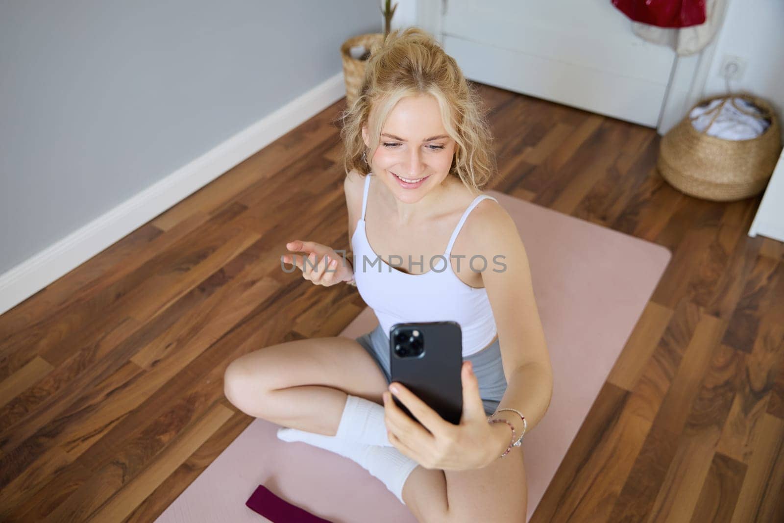 Young sporty fitness trainer, yoga instructor does live stream, takes selfies on smartphone while sitting on rubber mat at home, recording her workout training session.