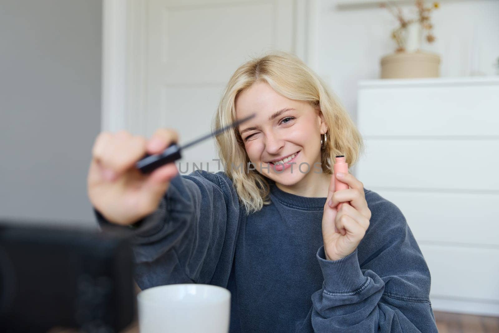 Portrait of young smiling woman in her room, recording video on camera, lifestyle vlog for social media, holding mascara, reviewing her makeup beauty products, showing how to use cosmetics.