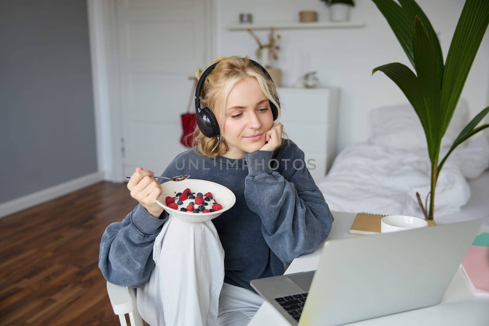 Portrait of happy young blond woman, sitting in a room, watching movie on laptop and eating healthy breakfast, drinking tea, resting on weekend.