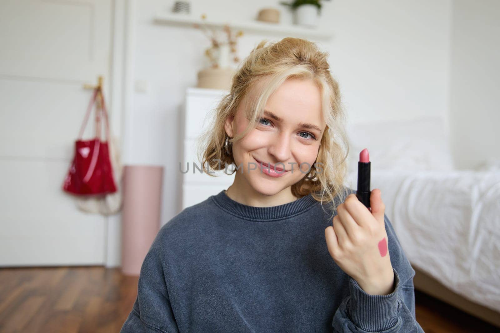 Portrait of smiling blond woman, recording video, online live stream about beauty products, content maker rates her makeup products, shows lipstick on camera.